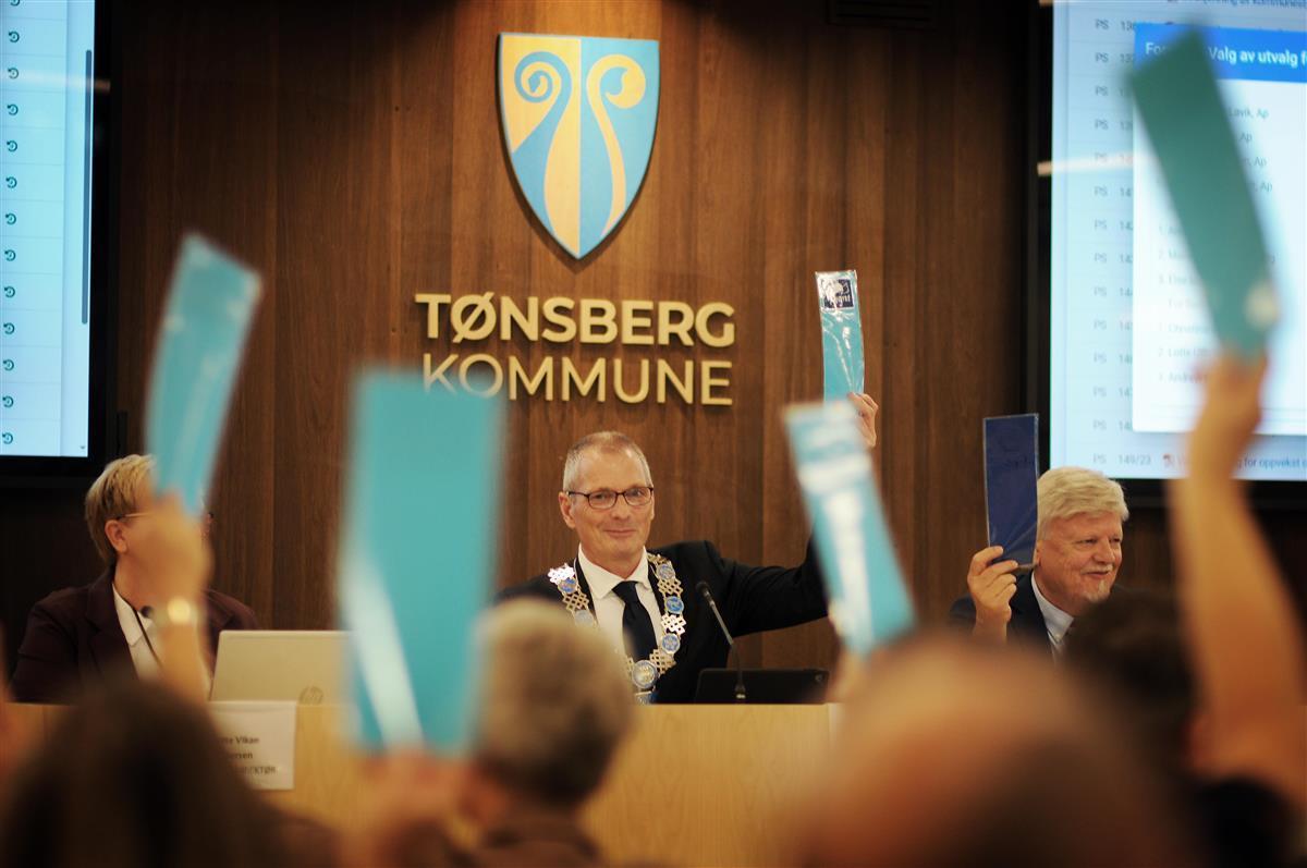 Ordføreren på podiet i kommunestyresalen og stemmesedler som løftes opp. - Klikk for stort bilde