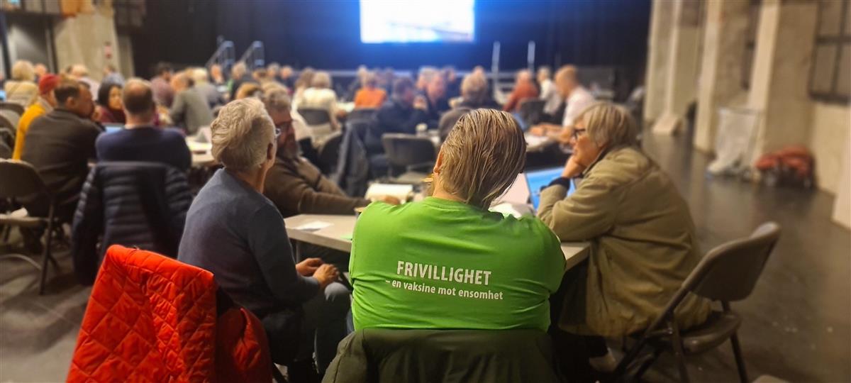 Folk som sitter sammen rundt flere bord. En mann sitter med ryggen til med en t-skjorte hvor det står at frivillighet er en vaksine mot ensomhet. - Klikk for stort bilde